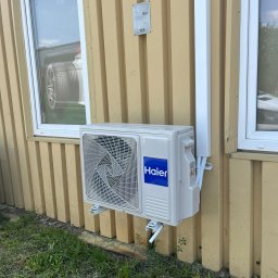 InstalSilesia - Zamontowany klimatyzator Haier na elewacji budynku z żółtymi panelami, widoczny fragment okna z odbiciem opony samochodowej. Instalacja przy ziemi, zarośnięta trawą.