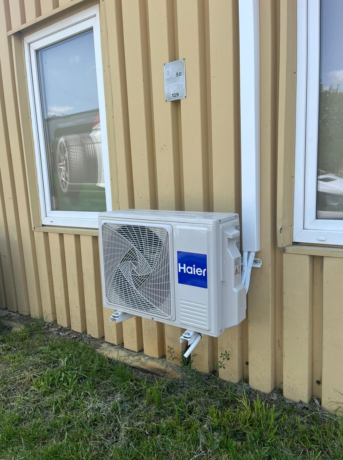 Zamontowany klimatyzator Haier na elewacji budynku z żółtymi panelami, widoczny fragment okna z odbiciem opony samochodowej. Instalacja przy ziemi, zarośnięta trawą.