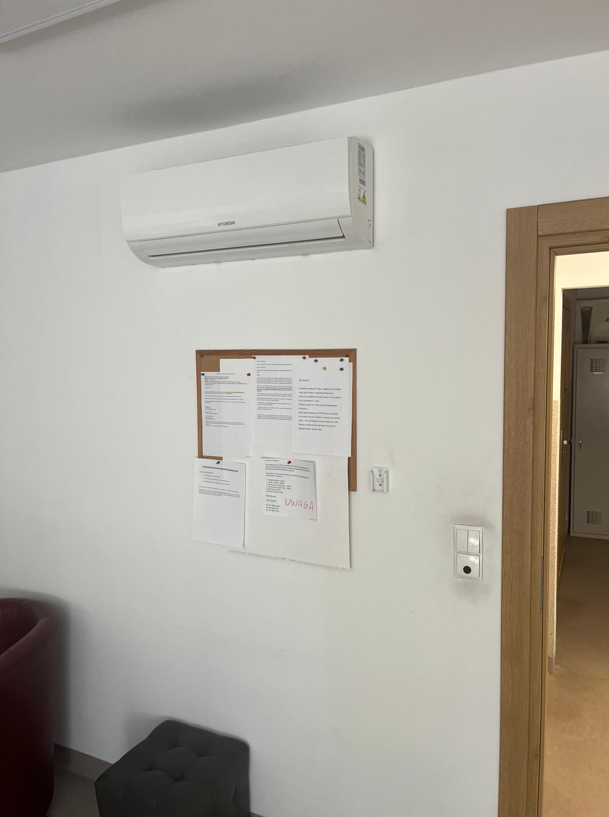 Biały klimatyzator Hyundai zamontowany na ścianie nad tablicą korkową z notatkami w jasnym pomieszczeniu. Widoczny fragment fotela i pufa.