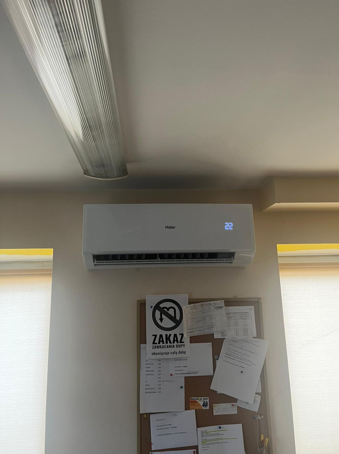 Biały klimatyzator Haier zamontowany na ścianie nad korkową tablicą z dokumentami, oświetlony długą lampą sufitową. Widoczny fragment okna z roletą.