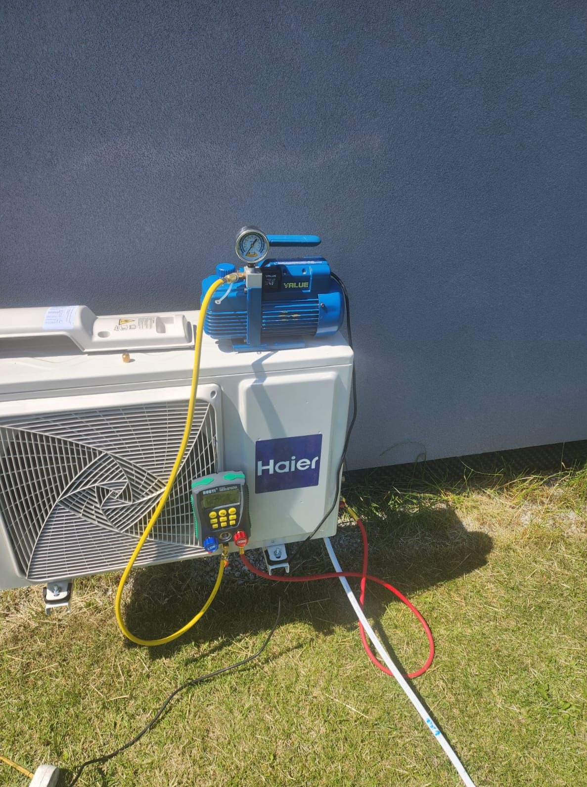 Błękitna pompa próżniowa VALUE i elektroniczny manifold Ruby's podłączone do białej jednostki zewnętrznej klimatyzacji Haier na tle szarej ściany i trawy.
