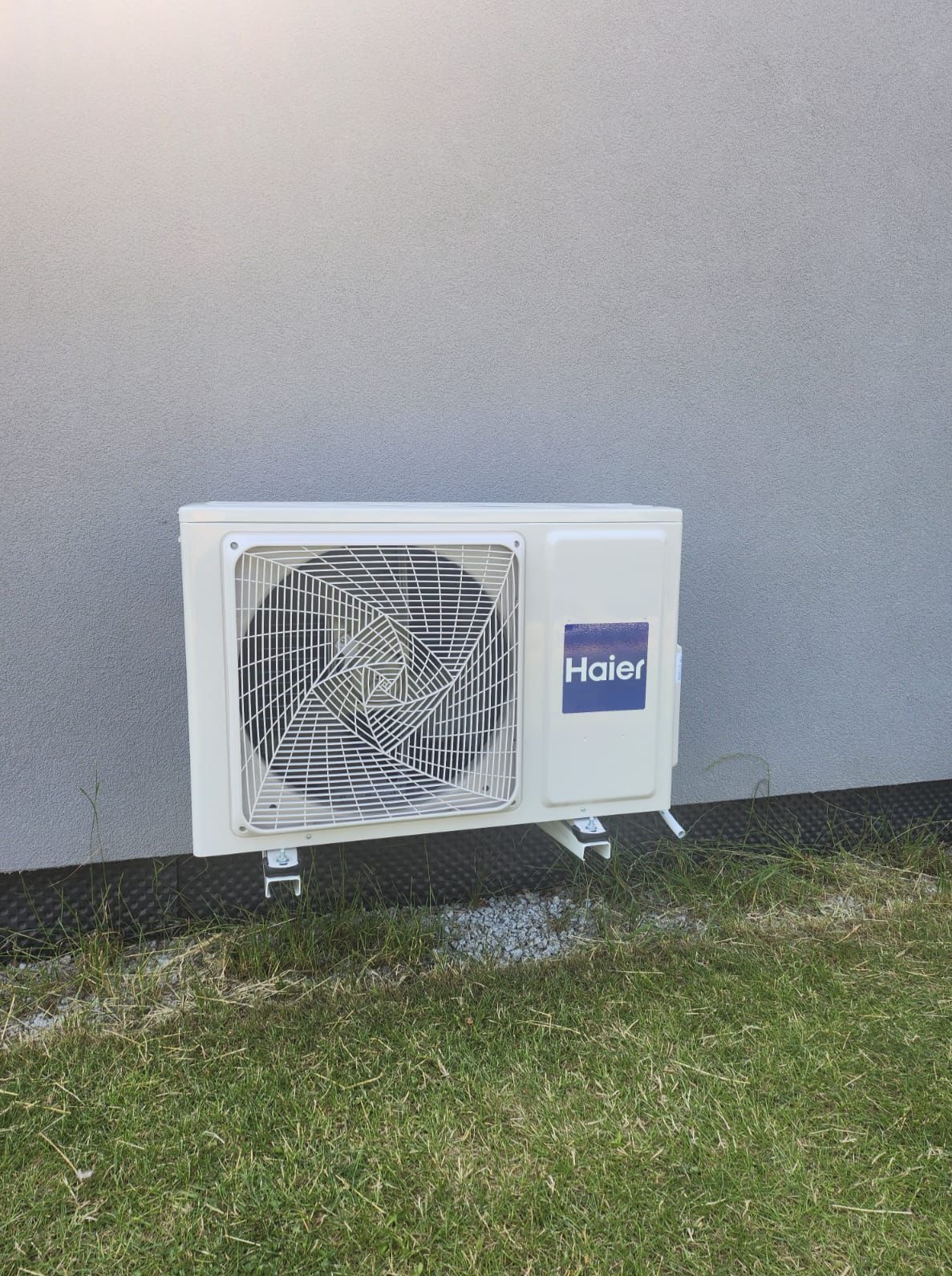 Zamontowana klimatyzacja Haier na ścianie budynku w Będzinie. Widoczna zewnętrzna jednostka klimatyzatora na tle trawy i elewacji.