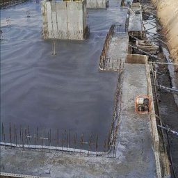 M-Bud Przemysłówka - Wylany fundament z wystającymi prętami zbrojeniowymi i betonowymi elementami szalunkowymi. W tle drzewa i wykop ziemny. Widoczny sprzęt budowlany.