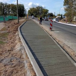 M-Bud Przemysłówka - Świeżo wylany, szary chodnik z betonową obwódką, na tle drogi i zieleni. Widoczni pracownicy i elementy zabezpieczeń drogowych.