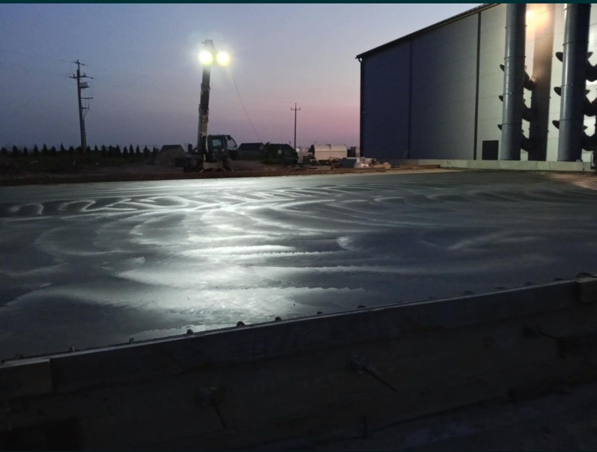 Świeżo wylana, mokra posadzka przemysłowa na zewnątrz, z widocznymi śladami po zacieraniu. W tle budynek magazynowy i maszyna budowlana oświetlająca teren.