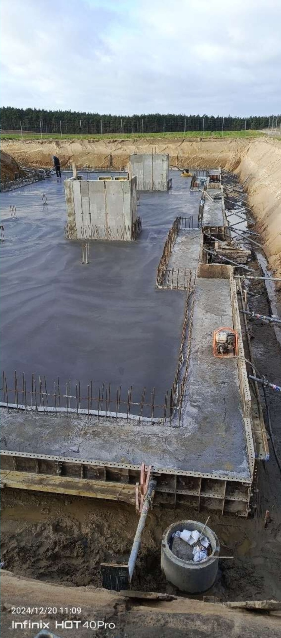 Wylany fundament z wystającymi prętami zbrojeniowymi i betonowymi elementami szalunkowymi. W tle drzewa i wykop ziemny. Widoczny sprzęt budowlany.