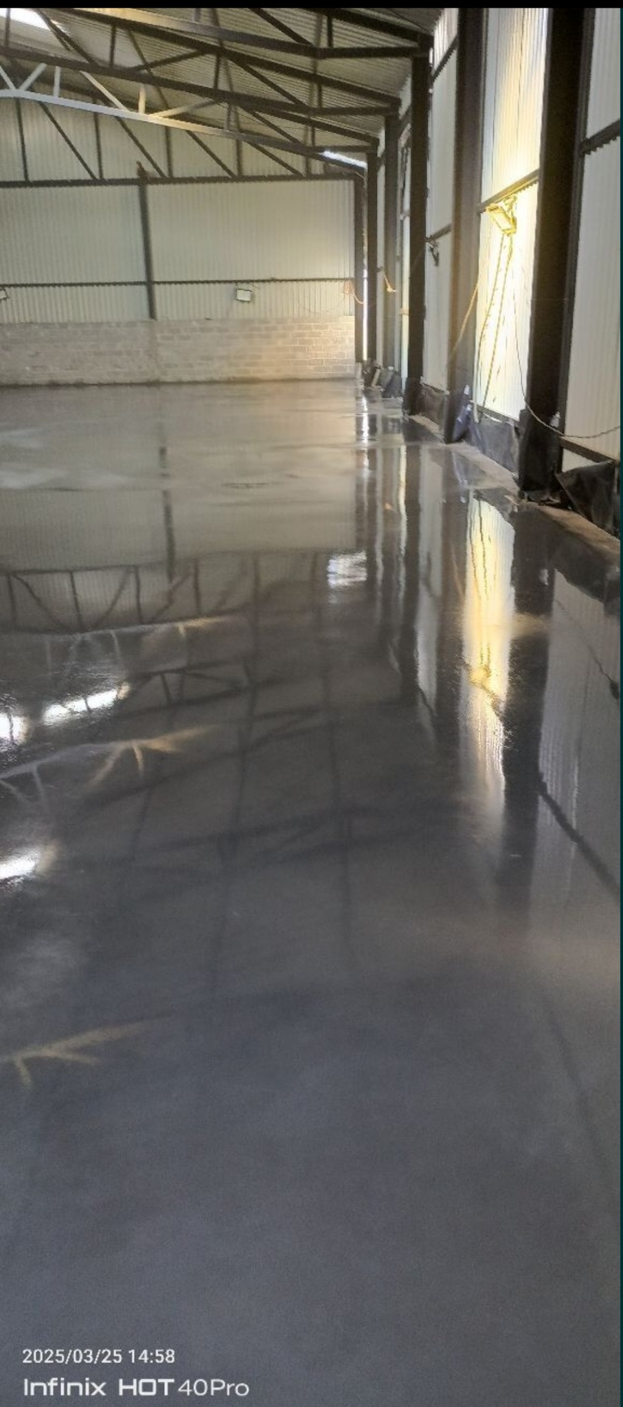 Gładka, szara posadzka przemysłowa w hali, z widoczną konstrukcją stalową dachu i ścianami z blachy trapezowej. Światło dzienne wpada przez okna.