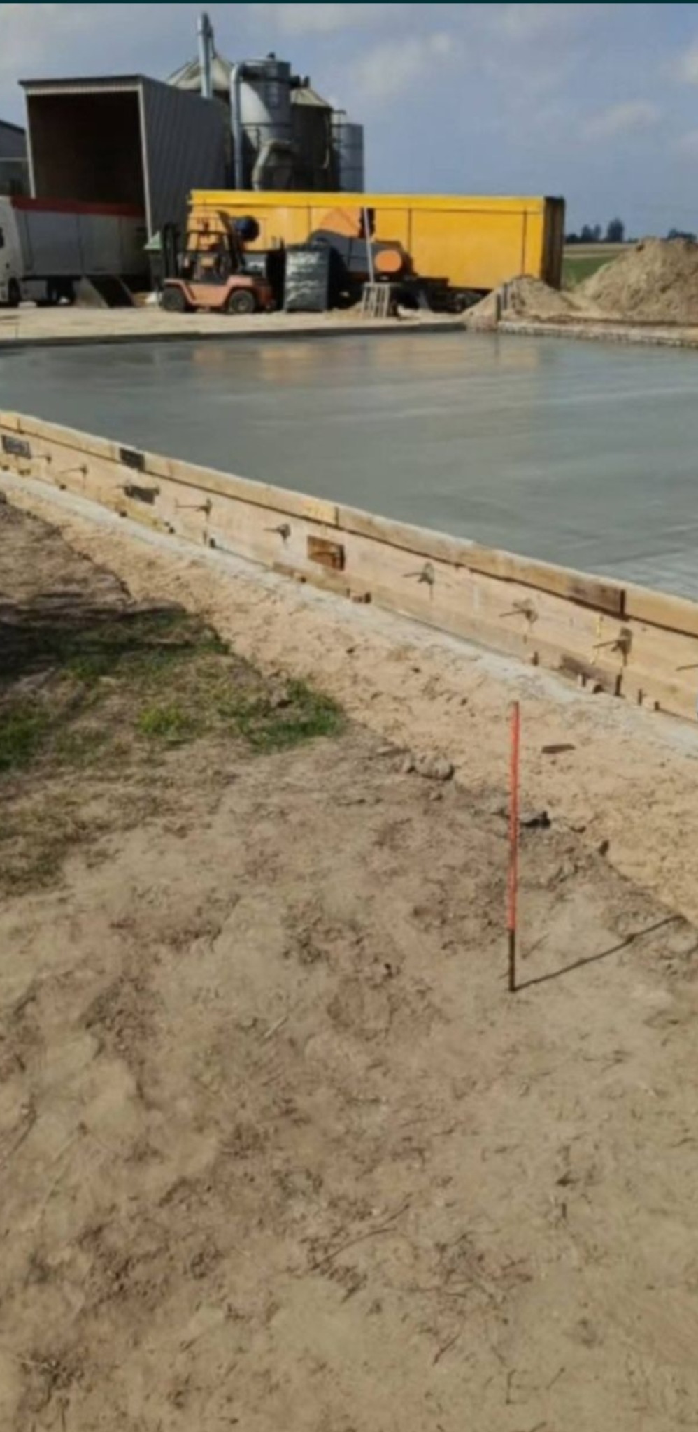 Wylewka betonowa na placu budowy, widoczne drewniane szalunki i sprzęt ciężki w tle. Świeżo wylany beton tworzy gładką powierzchnię.