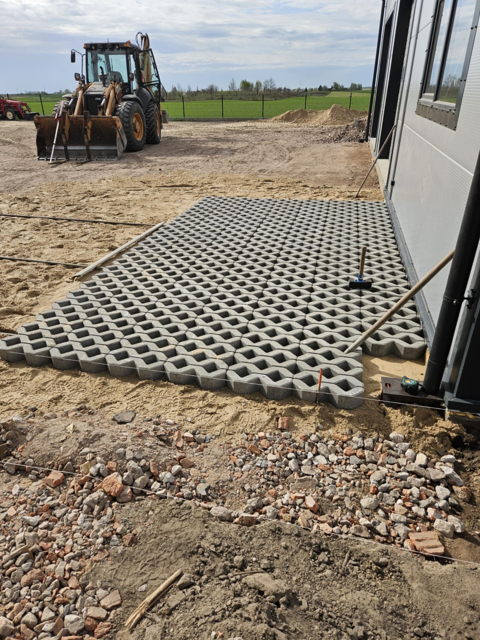 Układanie ażurowych płyt betonowych na piasku obok budynku. Widoczny traktor, narzędzia i fragment terenu z gruzem. Prace brukarskie w toku.