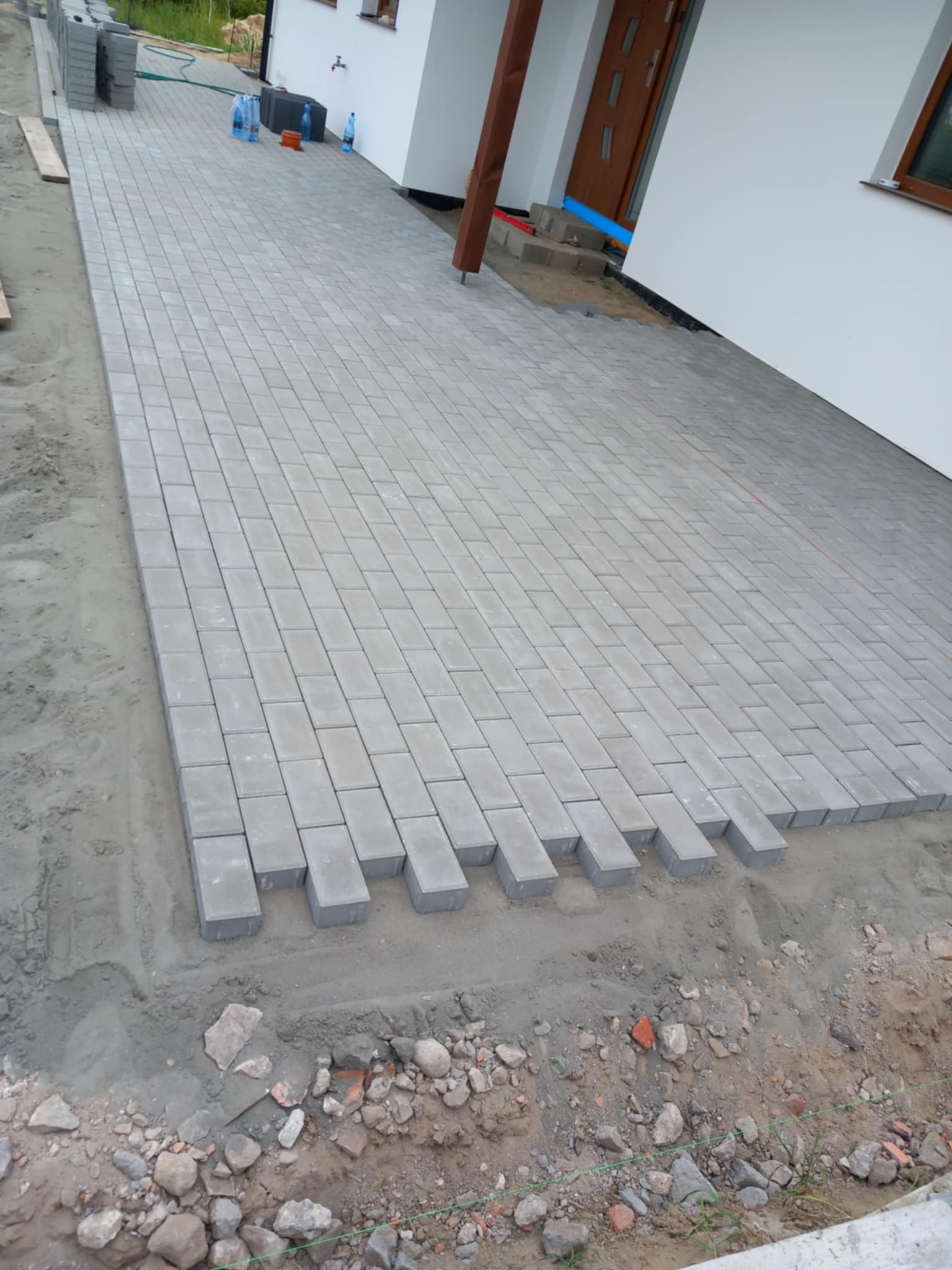 Montaż szarej kostki brukowej na posesji, widoczne niedokończone fragmenty i zapas kostki. Dom z białymi ścianami w tle.