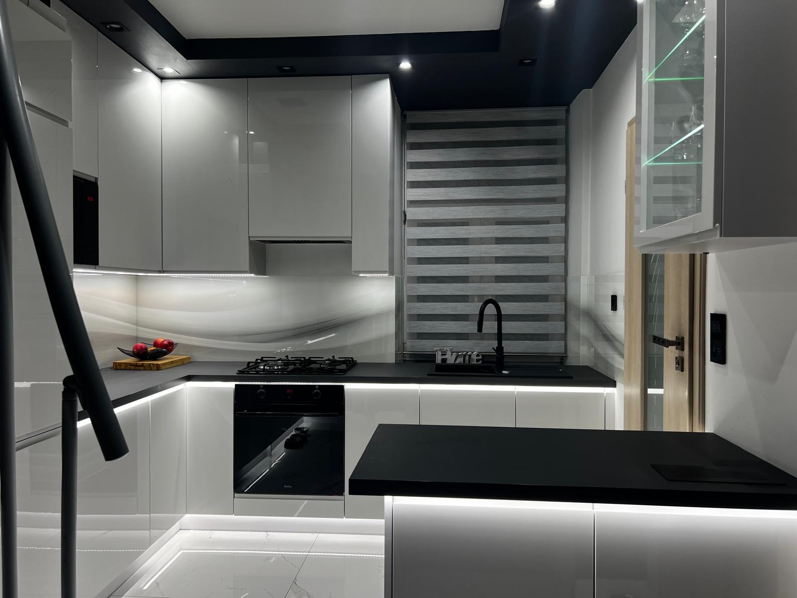 Nowoczesna, biała kuchnia z czarnymi blatami i oświetleniem LED pod szafkami. Widoczny fragment schodów, okno z żaluzjami i szklana szafka z zielonym podświetleniem.