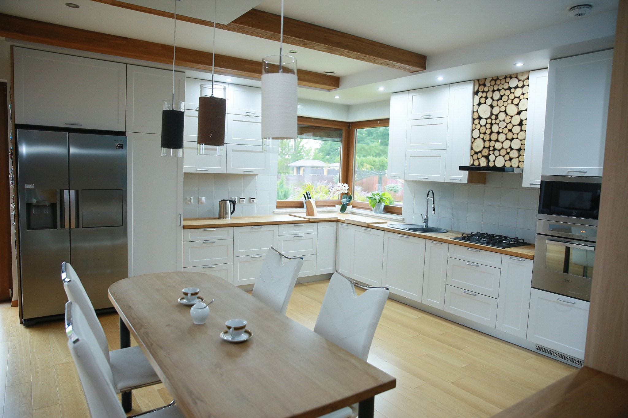 Jasna kuchnia z drewnianymi blatami, białymi frontami i akcentem z plastrów drewna nad kuchenką. Stół z zastawą i krzesłami w centrum.