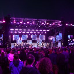 AGENCJA BRUSSA SC. - Scena z koncertu plenerowego, tłum widzów, orkiestra na podświetlonej scenie, fioletowe światła, duże ekrany po bokach sceny. Wydarzenie muzyczne w Bydgoszczy.