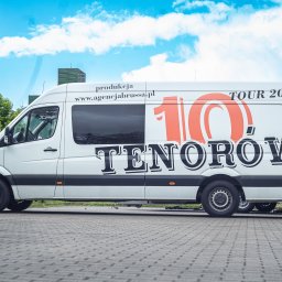 AGENCJA BRUSSA SC. - Biały van z logo '10 Tenorów TOUR 2025' i adresem agencjabrussa.pl, zaparkowany na brukowanej nawierzchni na tle zieleni i błękitnego nieba.