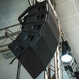 AGENCJA BRUSSA SC. - Profesjonalny system nagłośnieniowy line array, zawieszony na metalowej konstrukcji kratownicowej, oświetlony reflektorami. Widok z dołu, na tle sufitu.