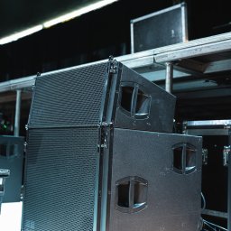 AGENCJA BRUSSA SC. - Zestaw profesjonalnych głośników estradowych, ustawionych jeden na drugim, z widocznymi kablami i elementami konstrukcji scenicznej w tle.