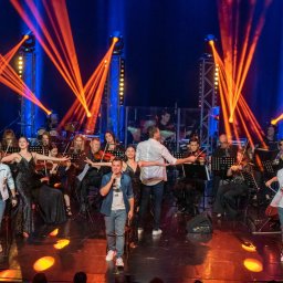 AGENCJA BRUSSA SC. - Zespół muzyczny i wokaliści na scenie podczas występu, oświetlenie sceniczne, orkiestra smyczkowa w tle. Koncert w pełnym składzie, dynamiczne światła.