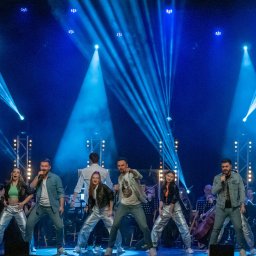 AGENCJA BRUSSA SC. - Zespół muzyczny na scenie w niebieskim oświetleniu, z orkiestrą w tle. Wokalistki i wokaliści w srebrnych spodniach śpiewają do mikrofonów.