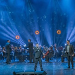 AGENCJA BRUSSA SC. - Wokalista w garniturze śpiewa na scenie z orkiestrą symfoniczną w tle, oświetlonej niebieskimi reflektorami podczas koncertu.