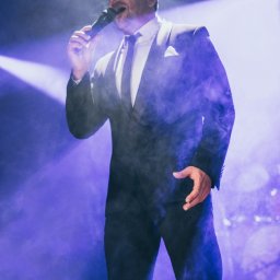 AGENCJA BRUSSA SC. - Wokalista w czarnym garniturze śpiewa do mikrofonu na scenie w fioletowym oświetleniu i dymie. Widoczny fragment sceny i publiczności.