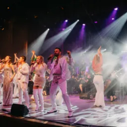 Zespół muzyczny na scenie w błyszczących strojach, oświetlony reflektorami podczas występu na żywo. W tle orkiestra i publiczność.