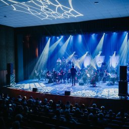 AGENCJA BRUSSA SC. - Orkiestra symfoniczna na scenie, oświetlona niebieskimi reflektorami, dyrygent w centrum, widownia w tle. Dekoracja sufitu z motywem pajęczyny.