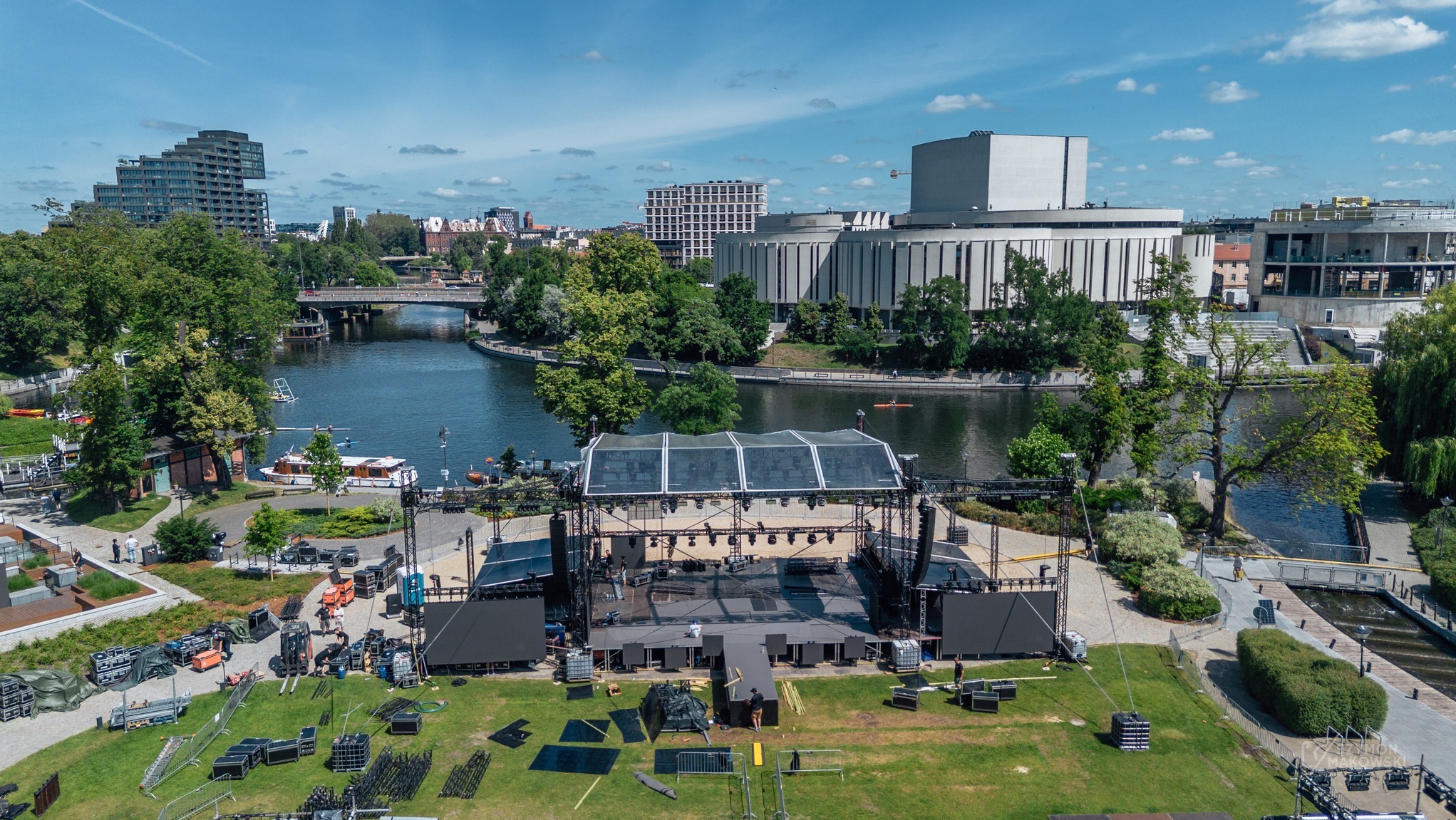 Scena plenerowa w Wełnicy w trakcie montażu, widok z góry. W tle rzeka i budynki. Sprzęt nagłośnieniowy i oświetleniowy rozstawiony na trawie.