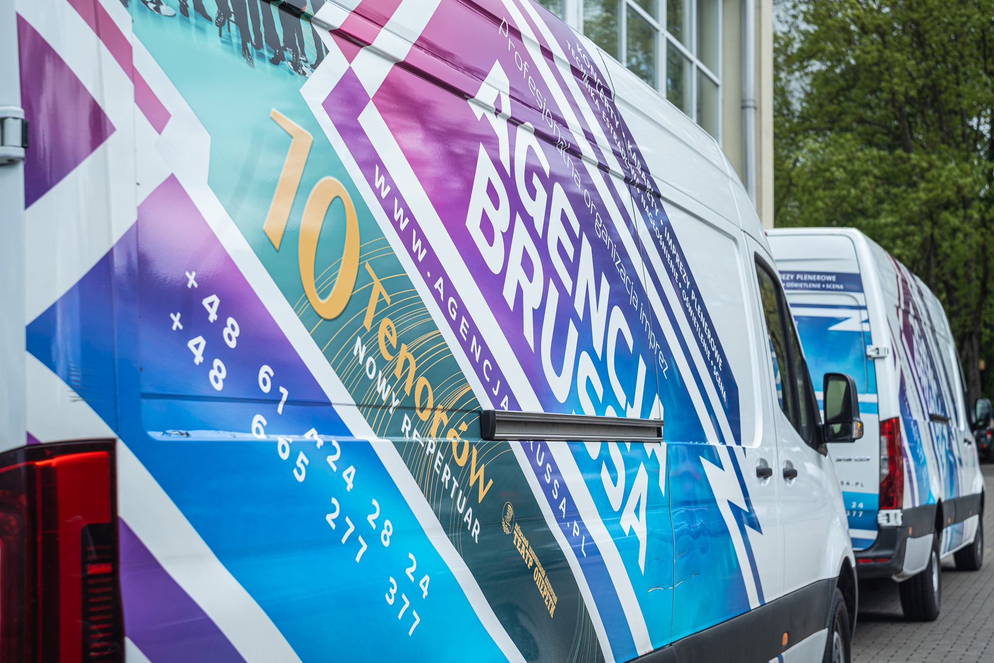 Flota białych busów Agencji Brussa z grafiką informującą o organizacji imprez, koncertów i nagłośnieniu. Widoczne numery kontaktowe i strona www agencji.