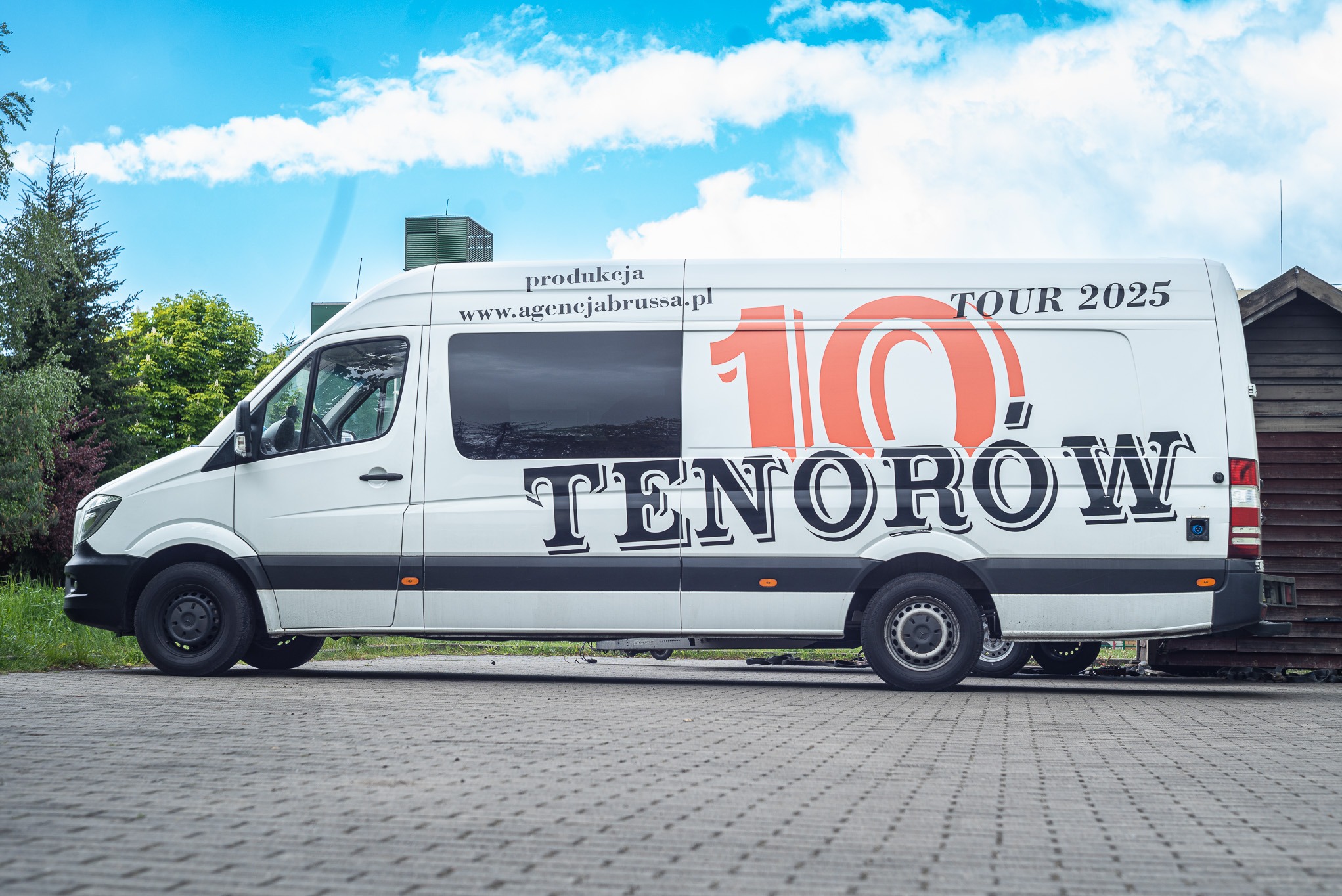 Biały van z logo '10 Tenorów TOUR 2025' i adresem agencjabrussa.pl, zaparkowany na brukowanej nawierzchni na tle zieleni i błękitnego nieba.