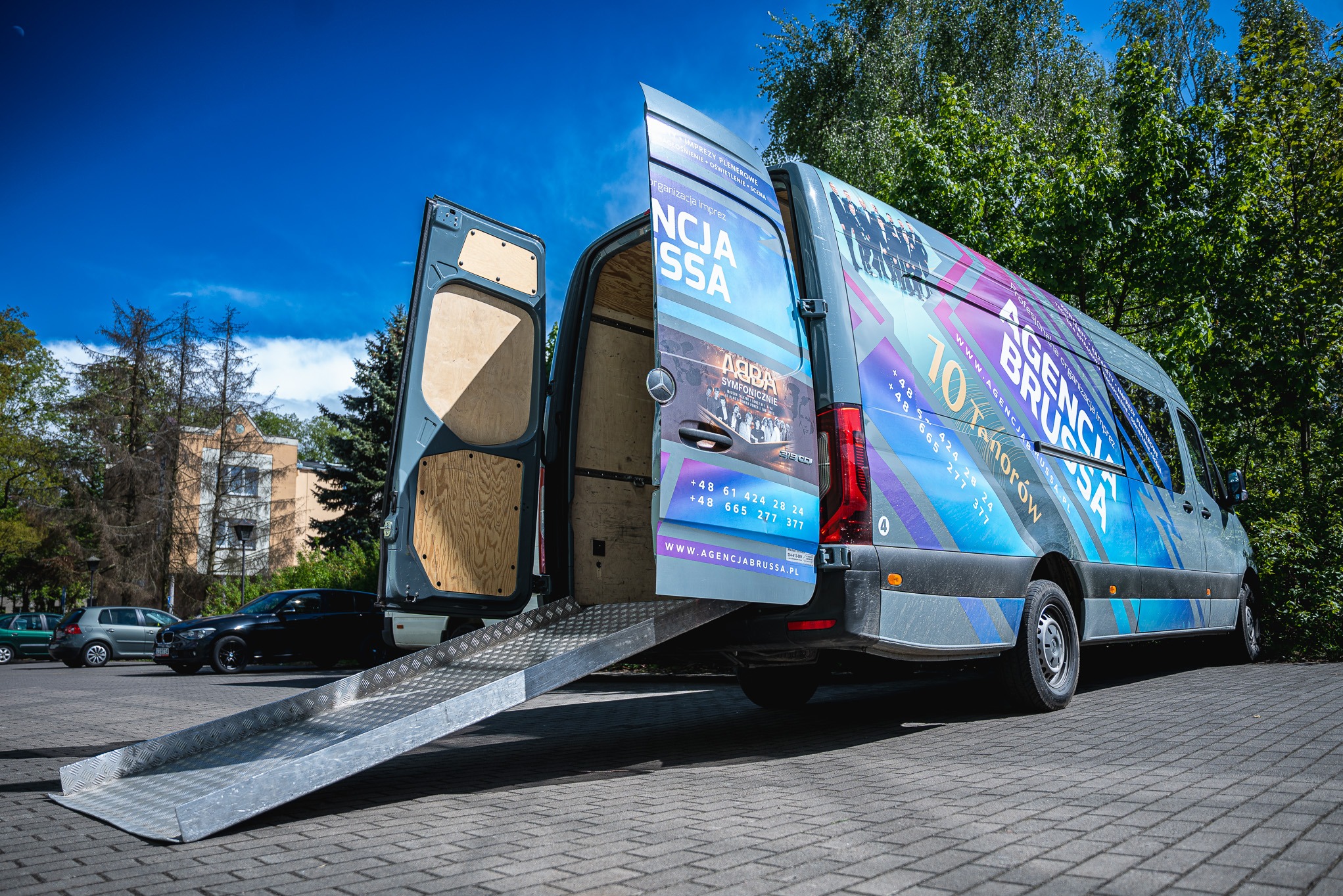 Van Agencji Eventowej Brussa z otwartymi drzwiami i rampą, reklama ABBA Symfonicznie i 10 Tenorów, błękitne niebo i zieleń drzew w tle.