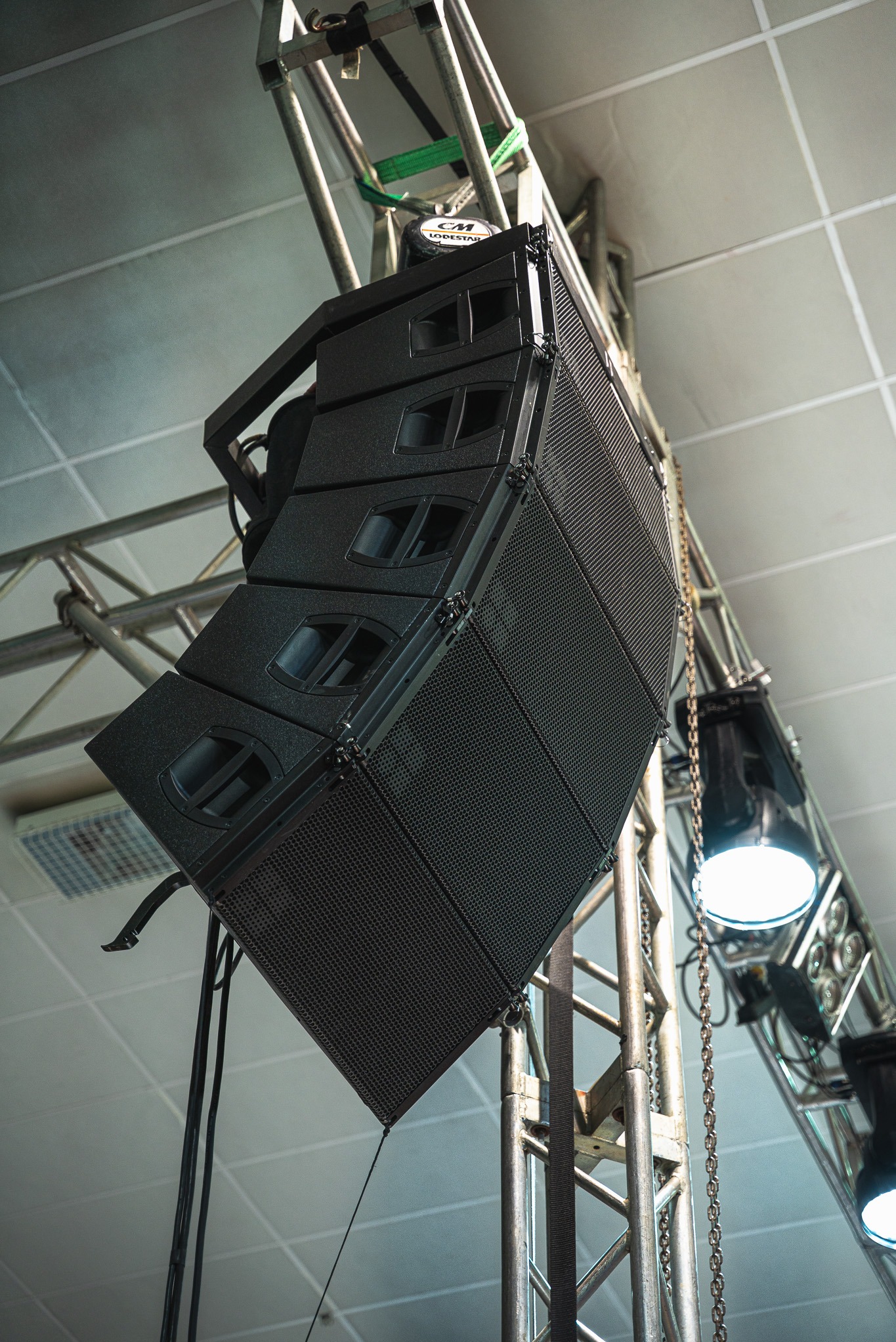 Profesjonalny system nagłośnieniowy line array, zawieszony na metalowej konstrukcji kratownicowej, oświetlony reflektorami. Widok z dołu, na tle sufitu.