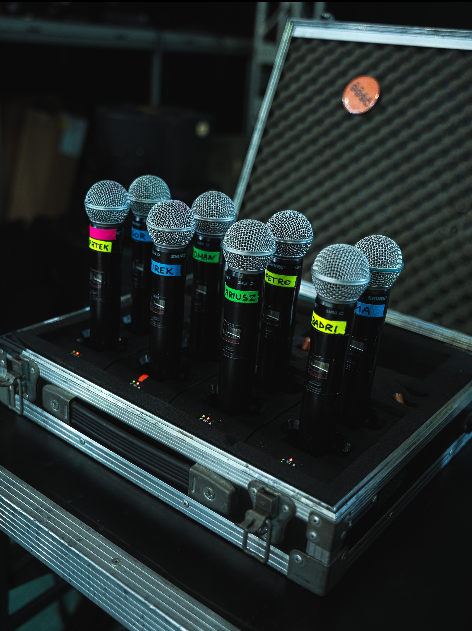 Zestaw mikrofonów Shure SM58 w metalowej walizce z piankowym wnętrzem, każdy oznaczony kolorową taśmą z imieniem, widoczny pomarańczowy znaczek na wieku walizki.