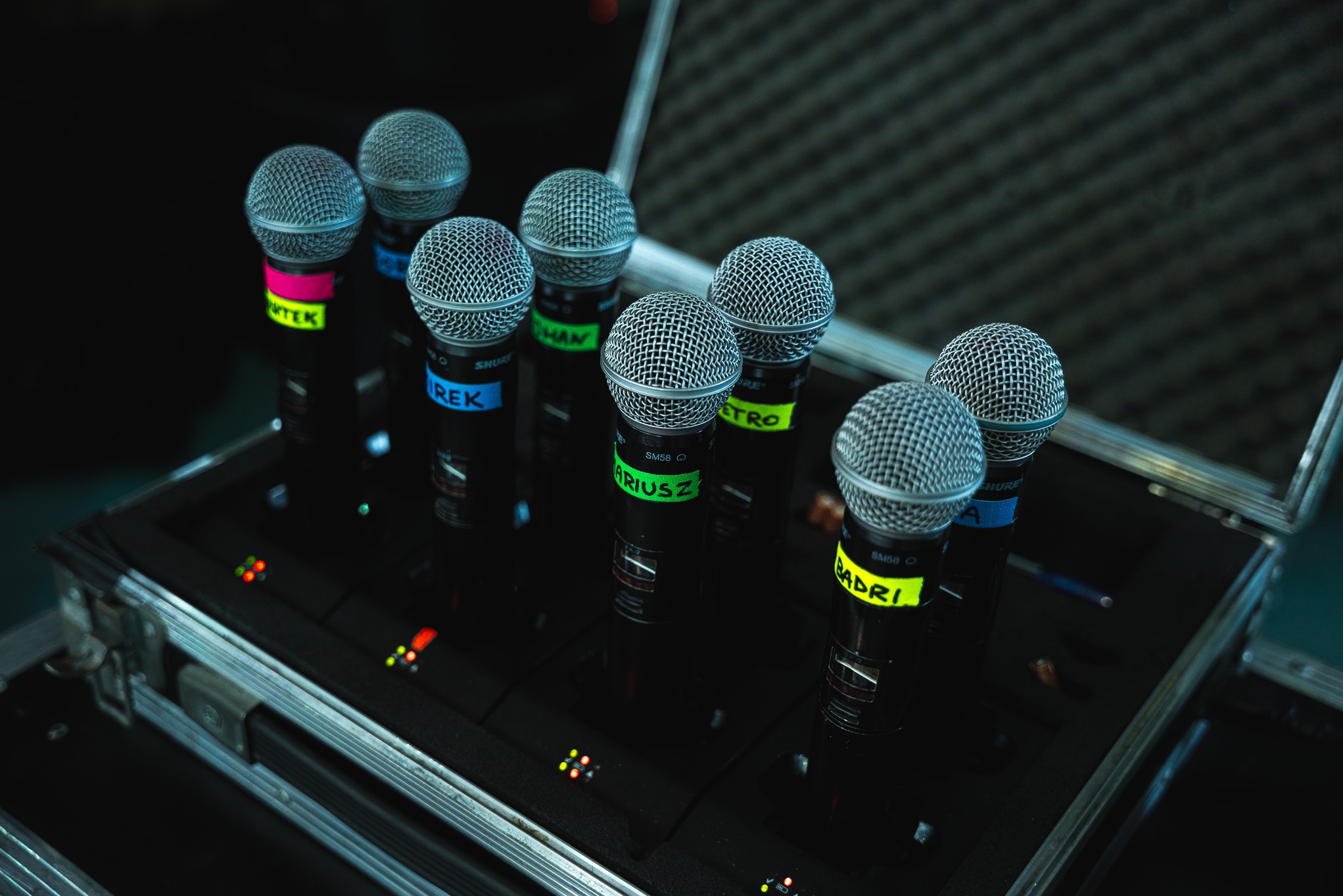Zestaw ośmiu mikrofonów Shure SM58 z kolorowymi oznaczeniami nazw, umieszczonych w czarnym, metalowym case'ie. Widoczne diody sygnalizacyjne.