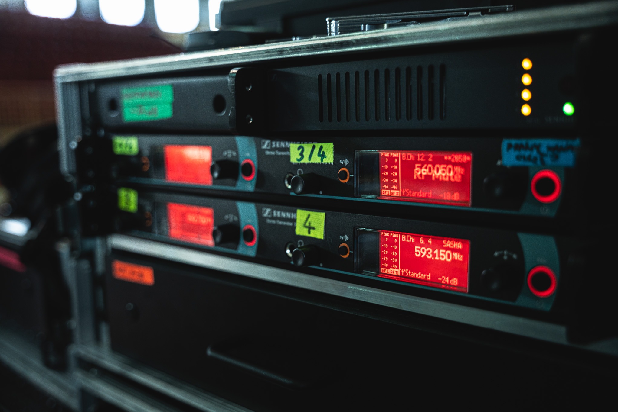 Sprzęt nagłośnieniowy Sennheiser w racku: odbiorniki bezprzewodowe z wyświetlaczami wskazującymi częstotliwość. Widoczne naklejki z numeracją kanałów.