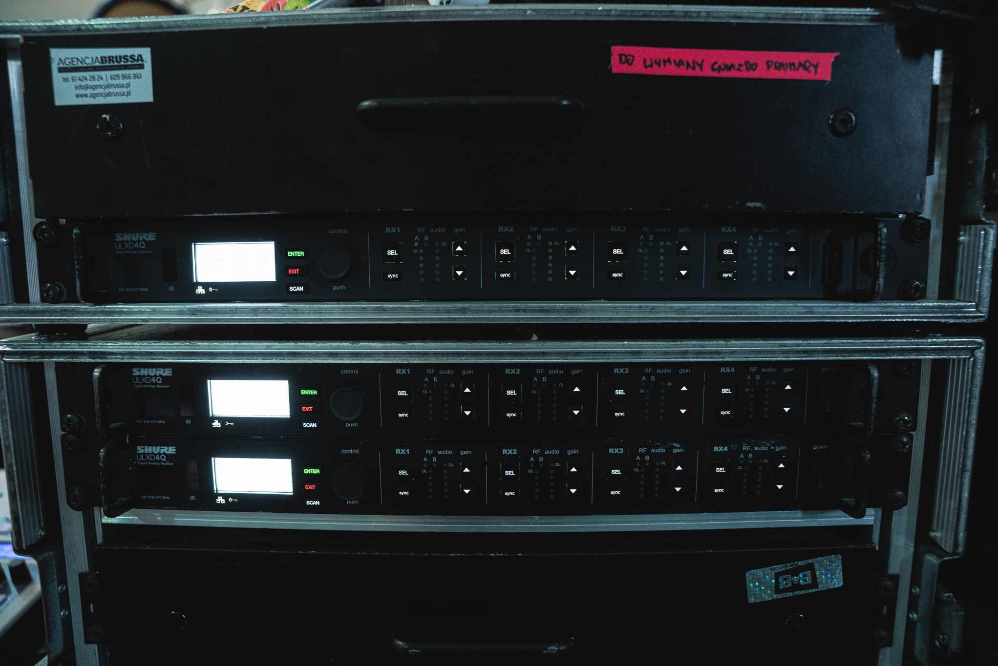 Sprzęt nagłośnieniowy Shure ULXD4Q w skrzyni transportowej. Widoczne odbiorniki bezprzewodowe z diodami i przyciskami. Etykieta 'DO WYMIANY GUMZDO PRIMARY'.