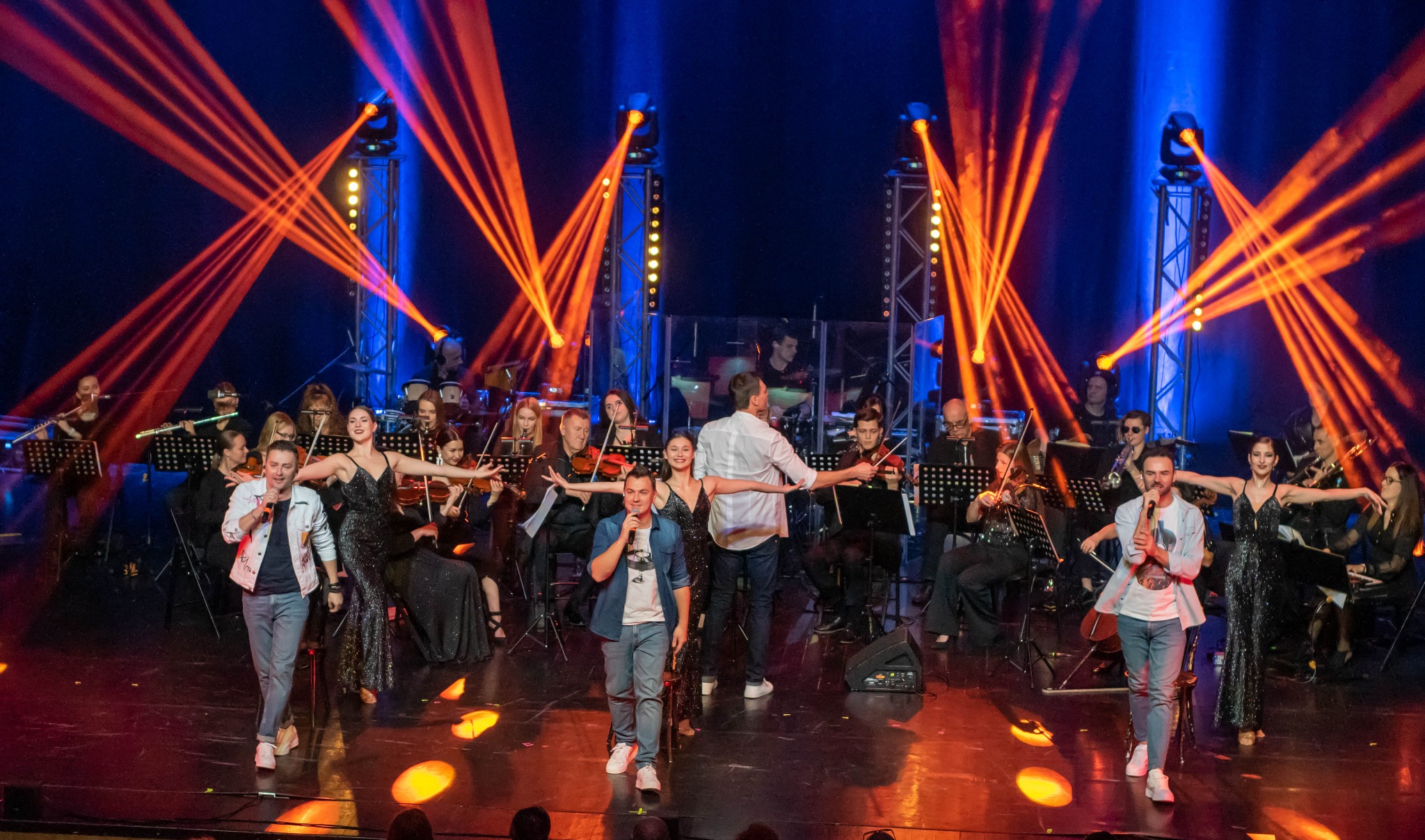 Zespół muzyczny i wokaliści na scenie podczas występu, oświetlenie sceniczne, orkiestra smyczkowa w tle. Koncert w pełnym składzie, dynamiczne światła.