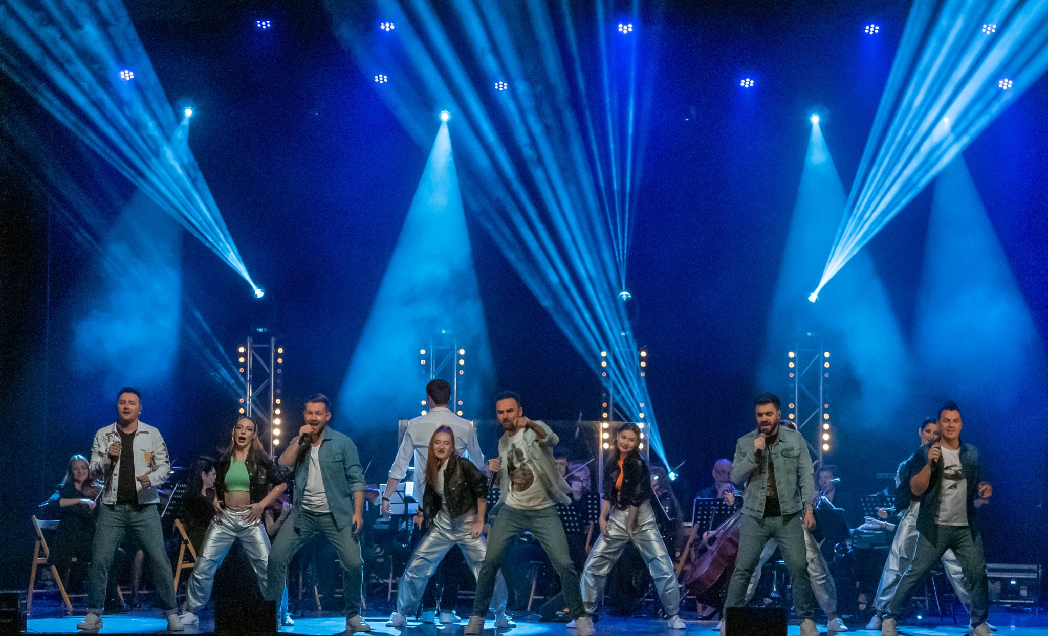 Zespół muzyczny na scenie w niebieskim oświetleniu, z orkiestrą w tle. Wokalistki i wokaliści w srebrnych spodniach śpiewają do mikrofonów.