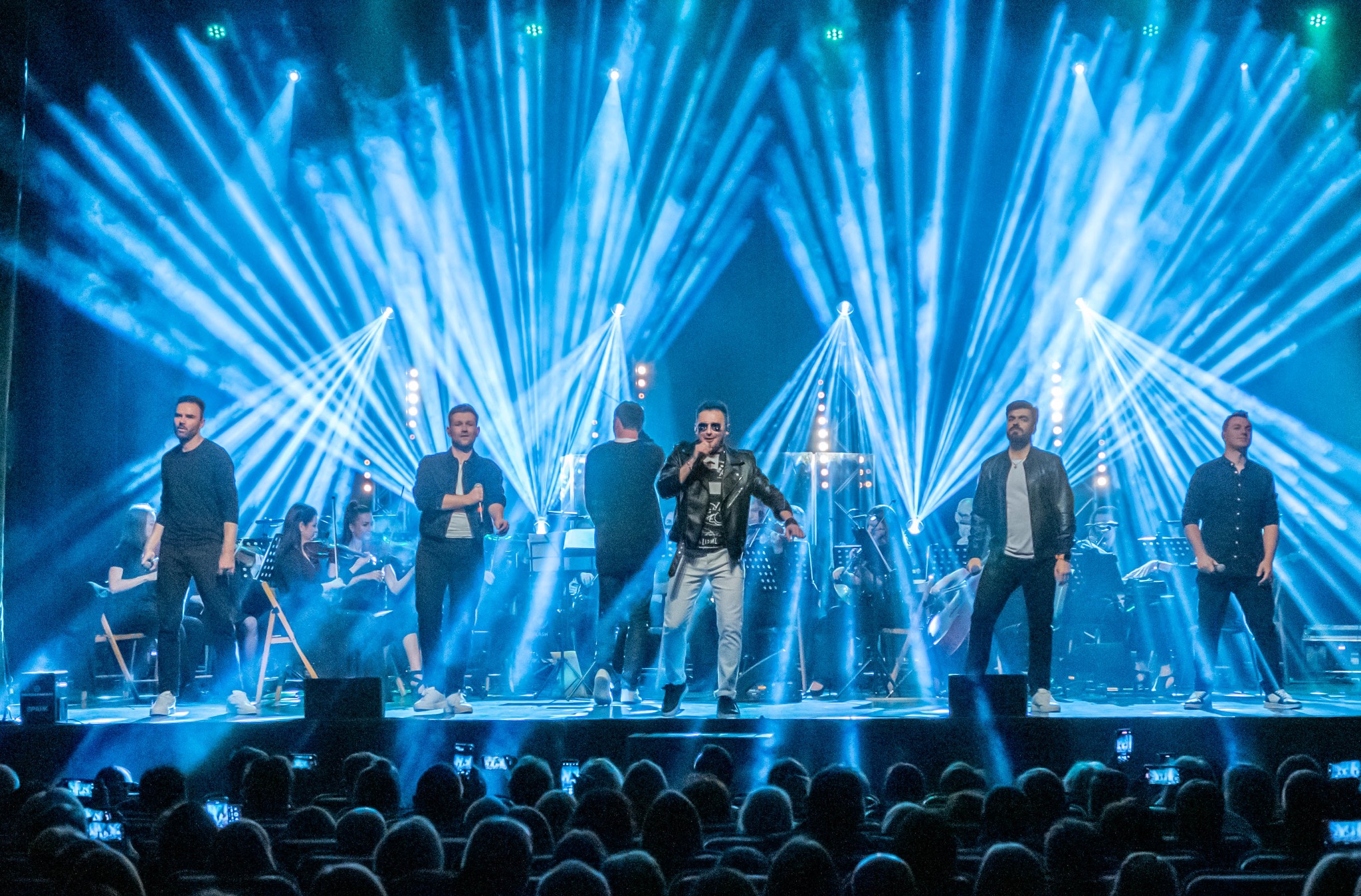 Zespół muzyczny na scenie podczas koncertu. Wokalista w skórzanej kurtce i okularach. Niebieskie oświetlenie sceniczne tworzy efektowne tło. Widownia filmuje występ.