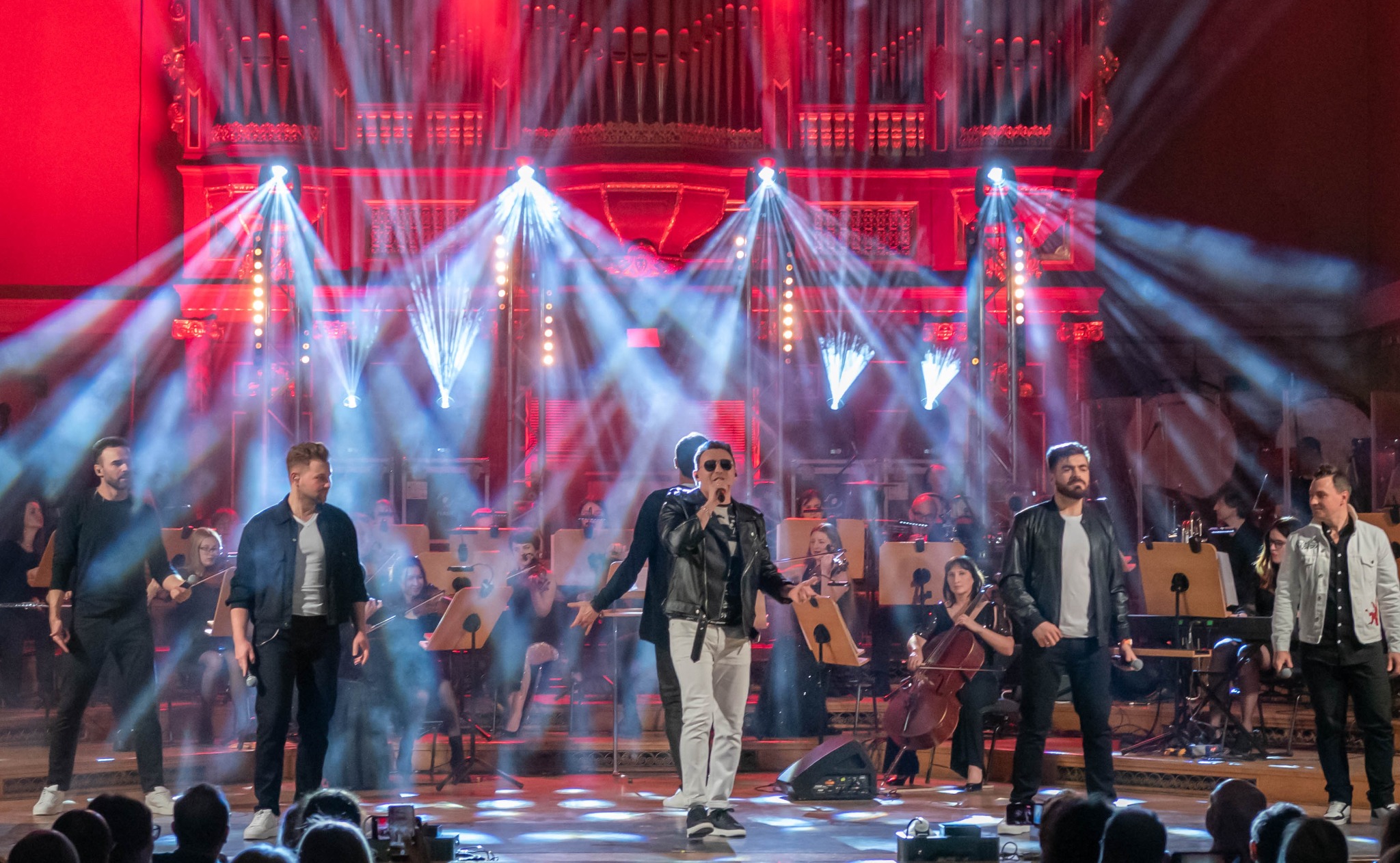 Zespół wokalny na scenie z orkiestrą w tle, oświetlony reflektorami. Wokalista w centrum, w skórzanej kurtce i białych spodniach, śpiewa do mikrofonu.