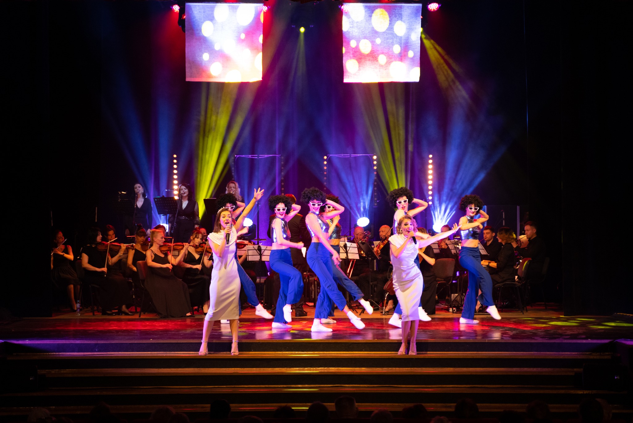 Scena z koncertu: wokalistki w białych sukienkach i tancerki w srebrnych topach i afro, orkiestra w tle, kolorowe światła i ekrany. Widok z dołu na widownię.