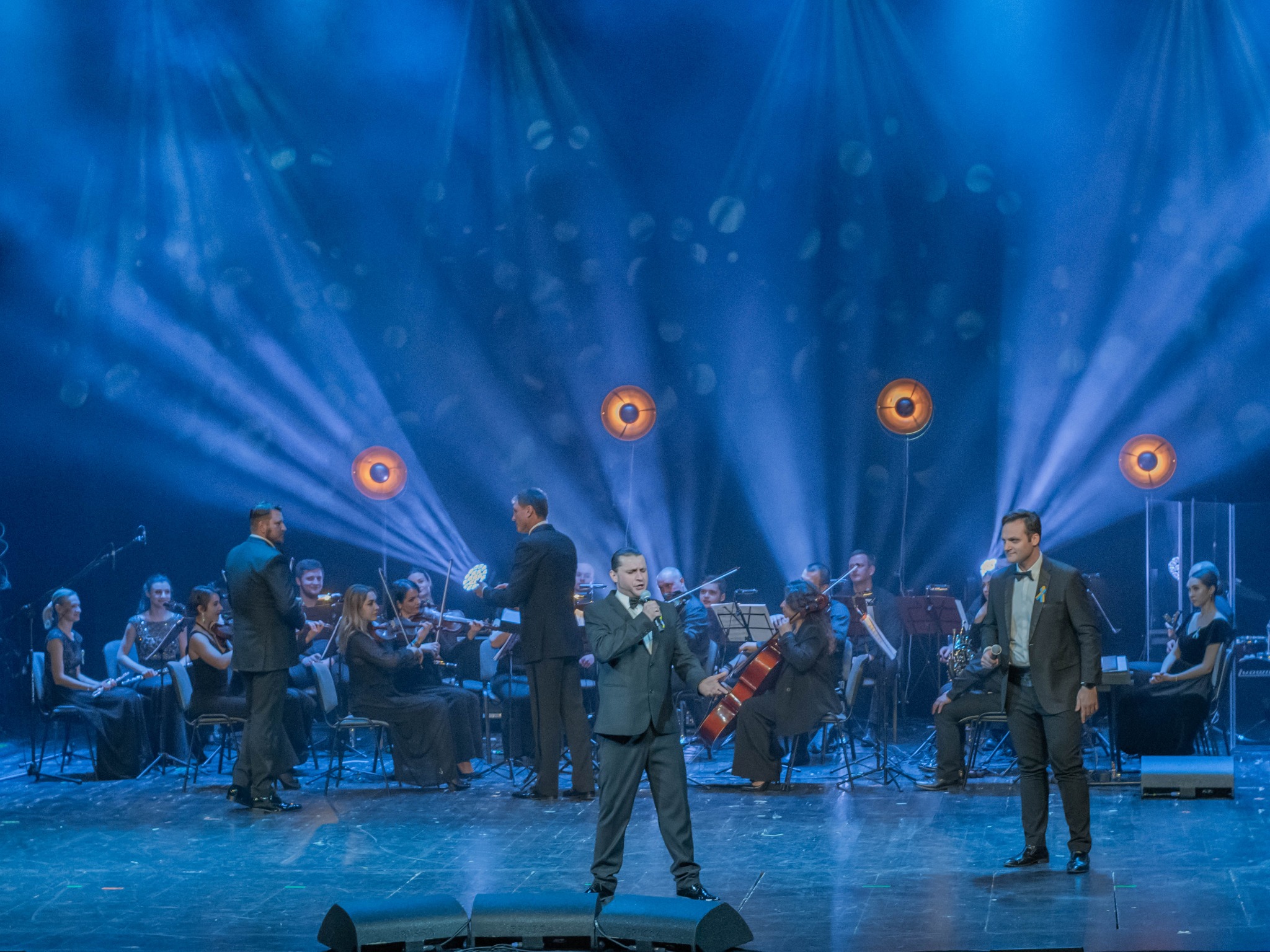 Wokalista w garniturze śpiewa na scenie z orkiestrą symfoniczną w tle, oświetlonej niebieskimi reflektorami podczas koncertu.