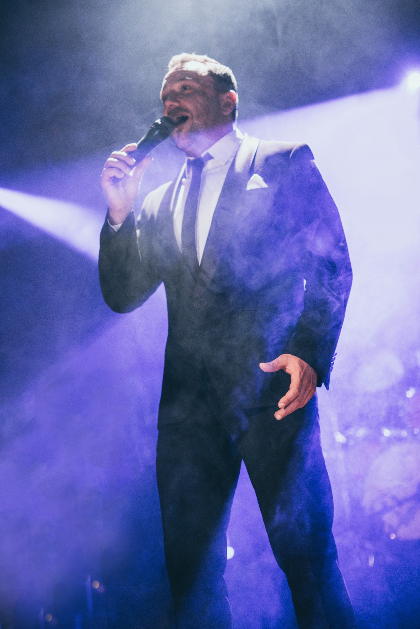 Wokalista w czarnym garniturze śpiewa do mikrofonu na scenie w fioletowym oświetleniu i dymie. Widoczny fragment sceny i publiczności.
