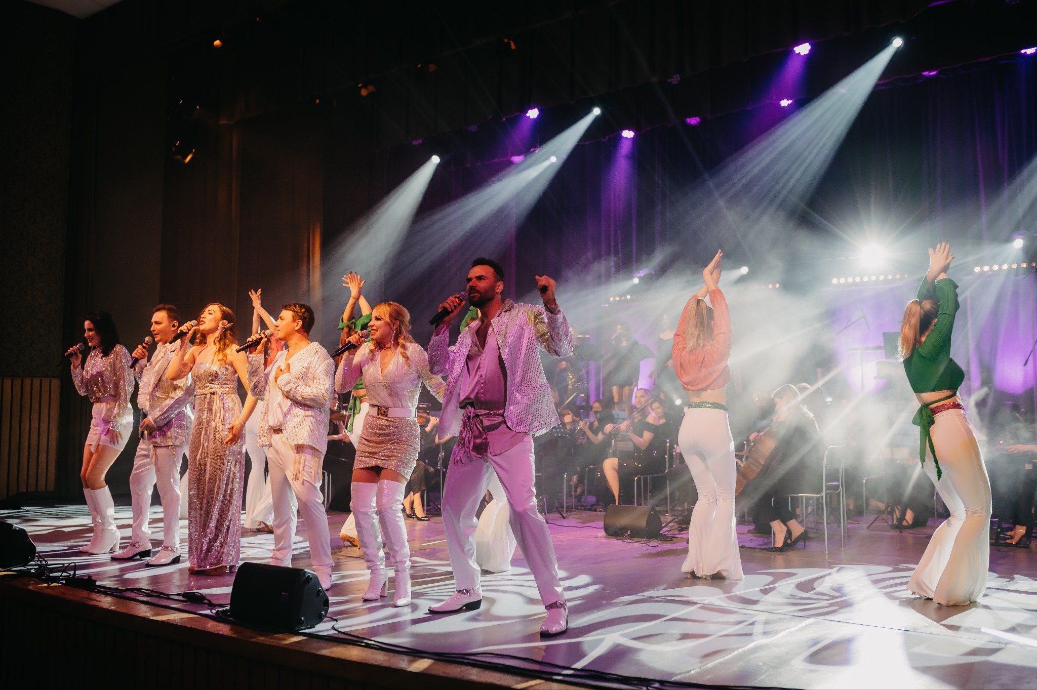 Zespół muzyczny na scenie w błyszczących strojach, oświetlony reflektorami podczas występu na żywo. W tle orkiestra i publiczność.
