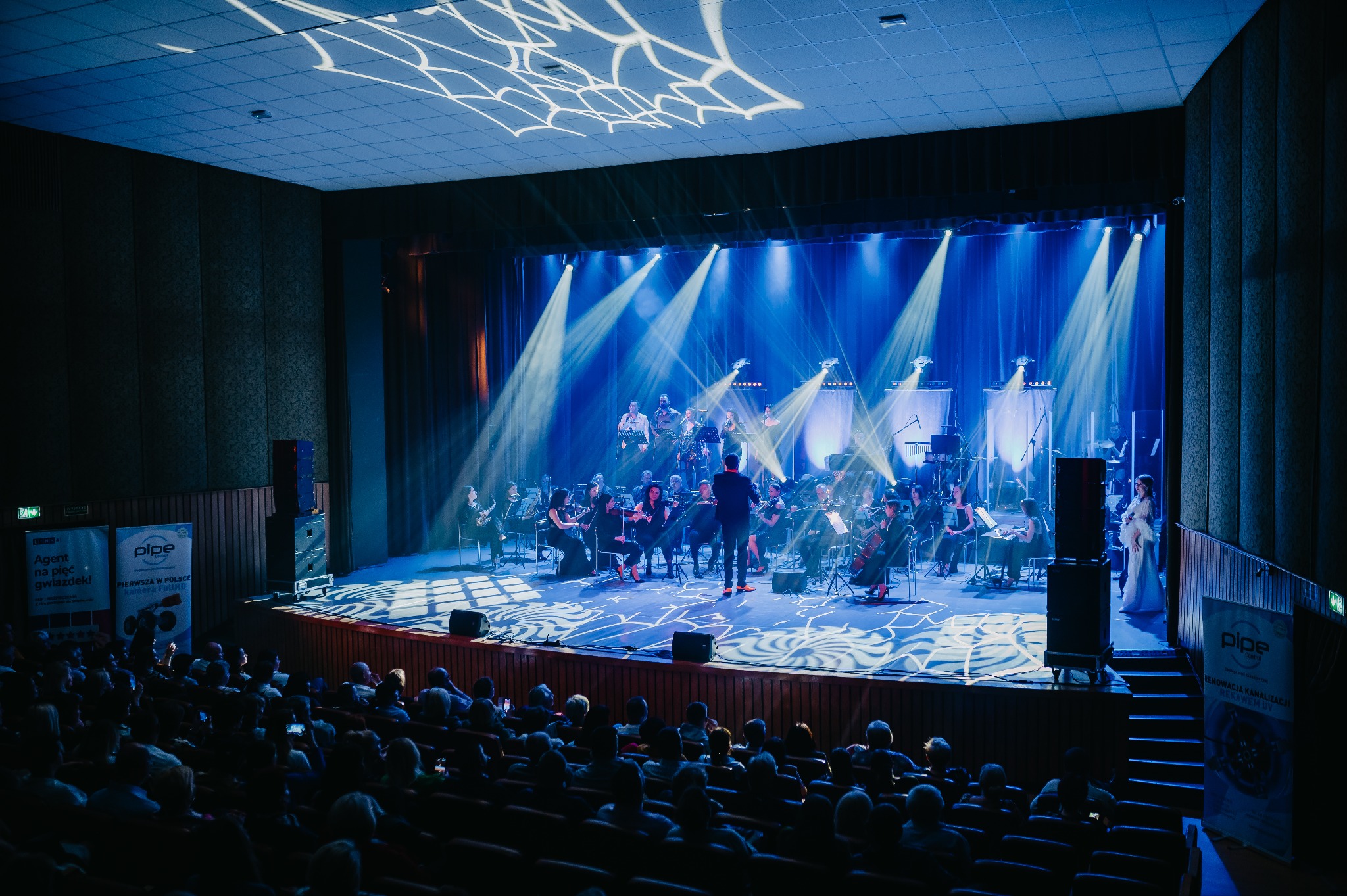Orkiestra symfoniczna na scenie, oświetlona niebieskimi reflektorami, dyrygent w centrum, widownia w tle. Dekoracja sufitu z motywem pajęczyny.