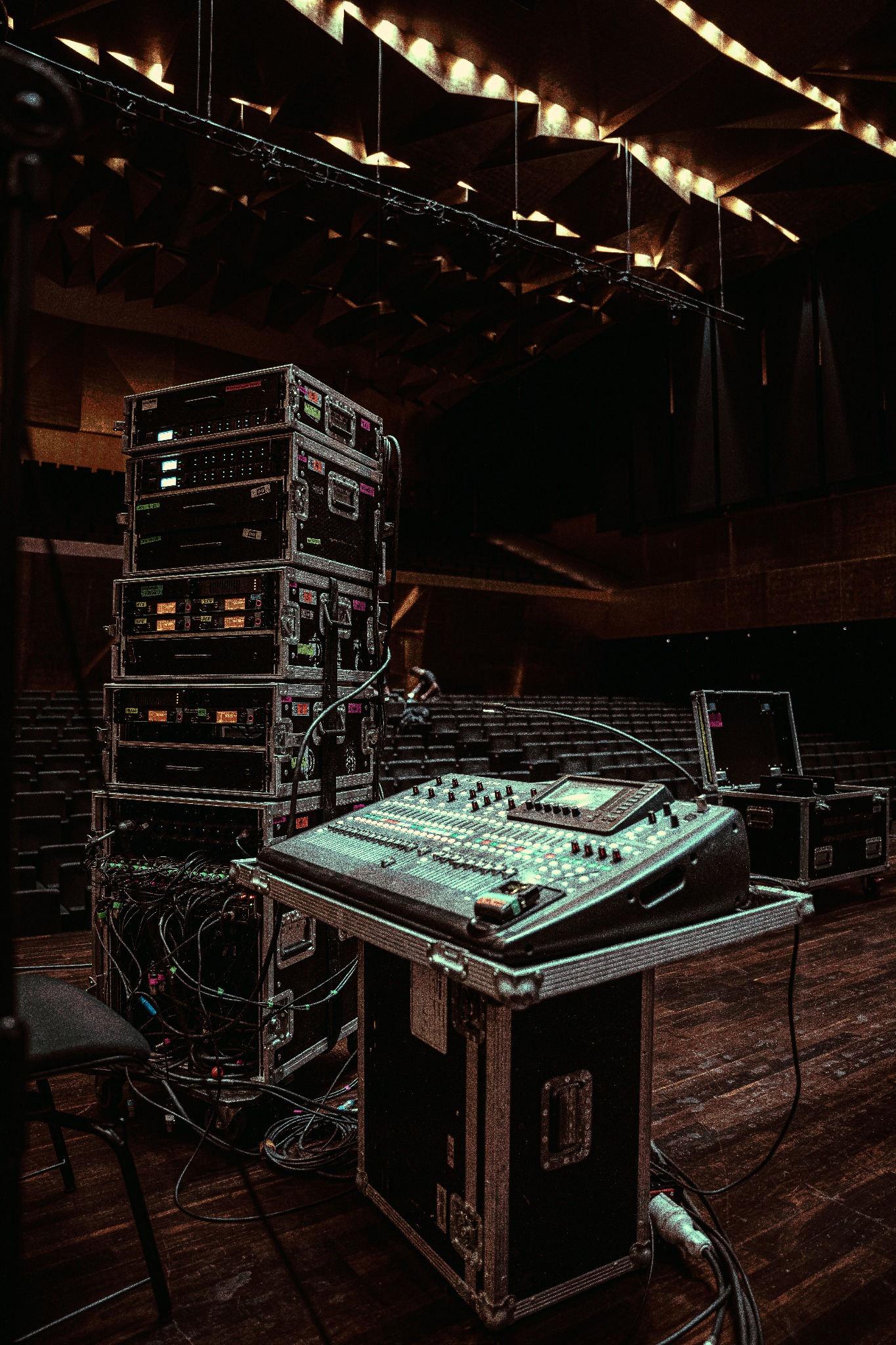 Sprzęt nagłośnieniowy: mikser i racki w case'ach na scenie. Oświetlenie sceniczne widoczne w tle, puste fotele na widowni. Profesjonalne nagłośnienie.