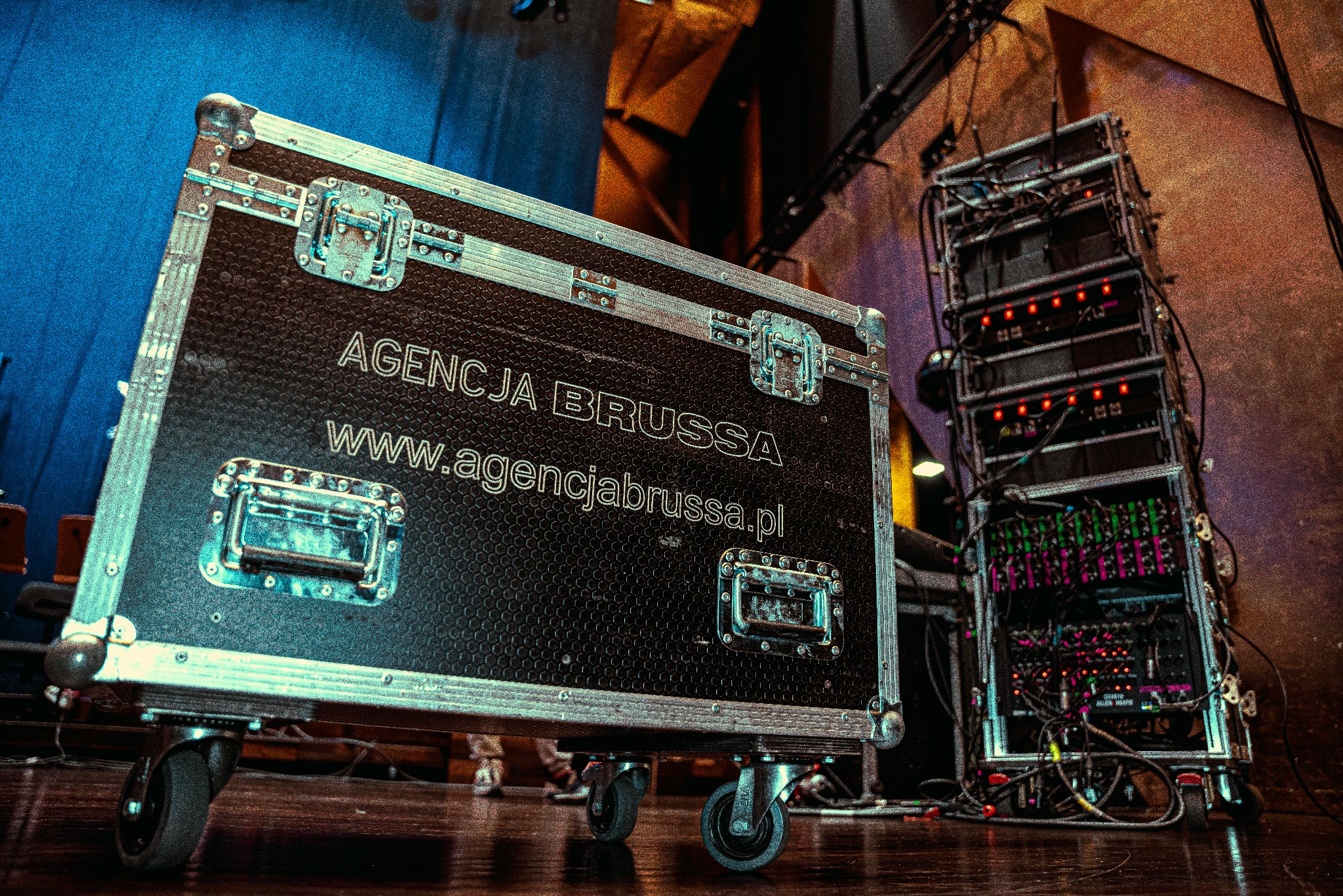 Skrzynia transportowa z nazwą Agencja Brussa i adresem strony internetowej, obok sprzęt nagłośnieniowy na scenie w ciepłych barwach, widok z dołu.