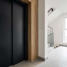 GB Foods Grzegorz Bandzarewicz - Nowoczesny hol wejściowy: czarna winda Otis, drewniane wykończenie i jasne płytki podłogowe. Minimalistyczne schody i okno w tle. Oświetlenie LED.