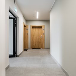 GB Foods Grzegorz Bandzarewicz - Korytarz w nowym budynku z szarymi płytkami podłogowymi, dwoje drzwi z numerami 388 i 389 oraz otwarte drzwi ewakuacyjne z czarną ramą.