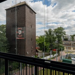 GB Foods Grzegorz Bandzarewicz - Widok z balkonu na ceglaną wieżę z logiem Boruta MC, drzewa, budynki i parking w tle. Ujęcie przez szklaną balustradę balkonu.