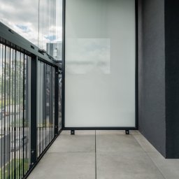 GB Foods Grzegorz Bandzarewicz - Nowoczesny balkon z szarymi płytkami, czarną balustradą z pionowymi szczebelkami i szklaną osłoną, z widokiem na zieleń i budynki w tle.